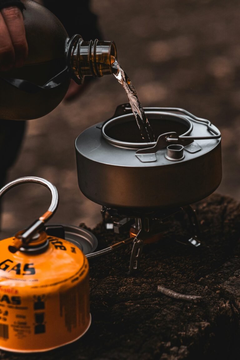 Outdoor camping scene with water being poured into a kettle on a portable gas stove.