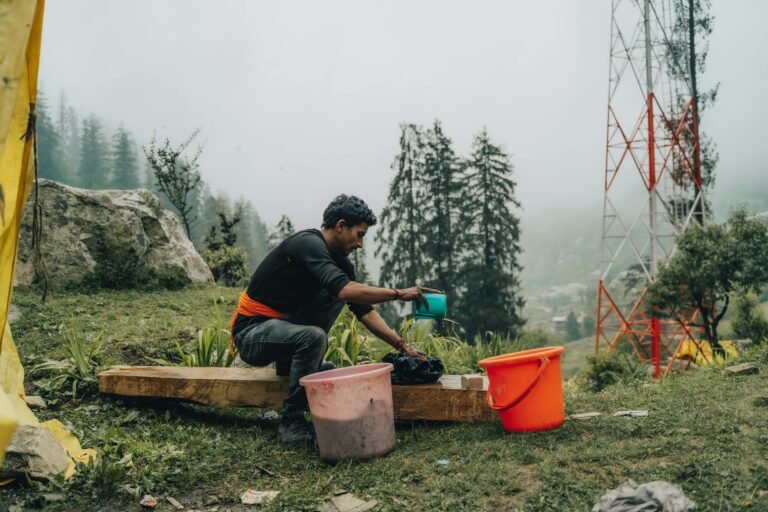 A man washes clothes outdoors in a rural area with misty trees and a utility tower in the background. ß Wohnmobil Waschmaschine