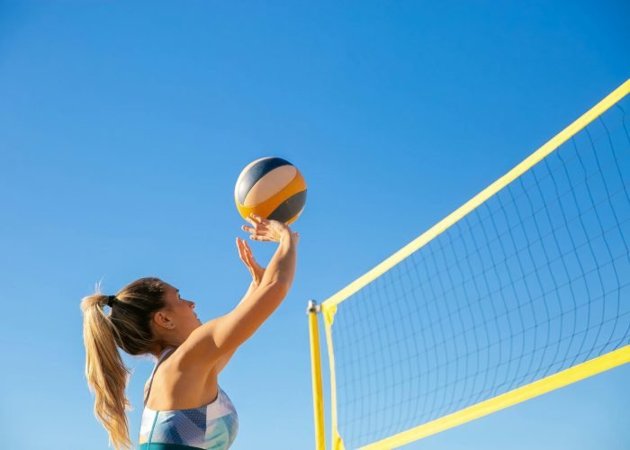 A woman plays volleyball on a sunny day, reaching to hit the ball over the net.